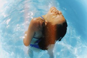 Person smiling with their head tilted back while standing in a sunlit blue swimming pool.