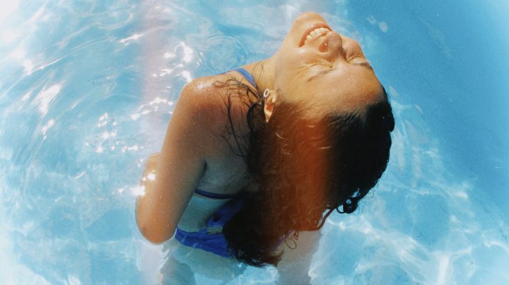 Person smiling with their head tilted back while standing in a sunlit blue swimming pool.