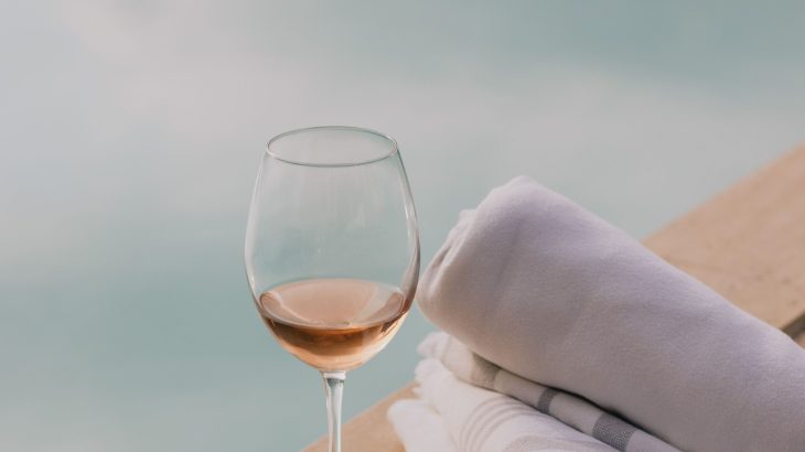 A partially filled glass of rosé wine sits next to neatly rolled white towels on a wooden pool deck, overlooking a tranquil swimming pool.