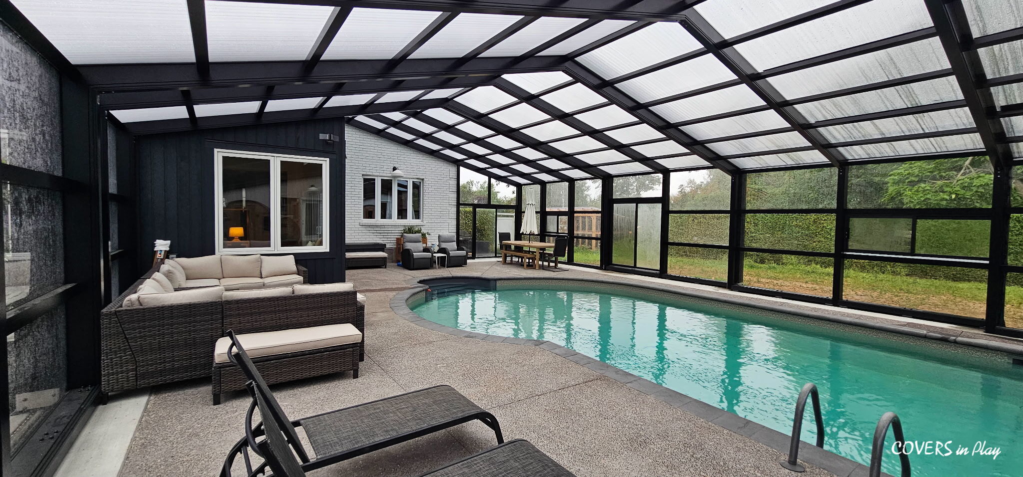 Enclosed patio with a curved indoor pool, outdoor furniture, a dining table, and large windows; clear roof and walls let in natural light.